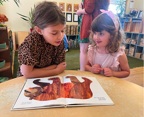 kindergarten-1 Older student reading a picture book with a younger child in a classroom.