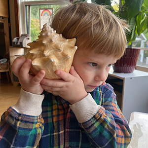 Child holding a large seashell to his ear in a classroom.